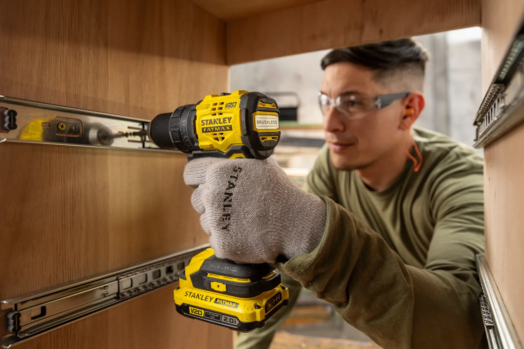 Fotografía de trabajador usando taladro inalámbrico STANLEY SBD714 dentro de gabinete de madera, con guantes y protección auditiva.