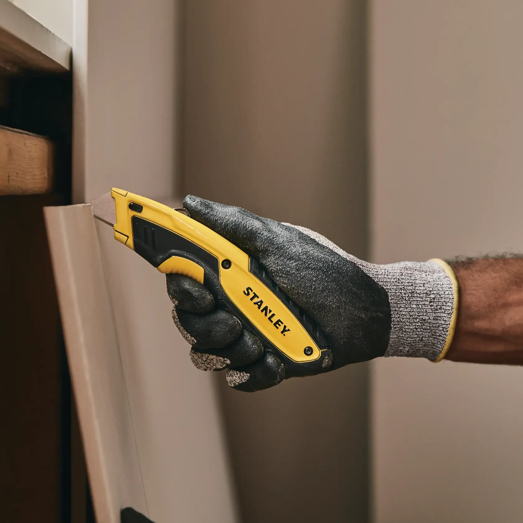 Estilete STANLEY STHT10479 com cabo amarelo e preto, mão com luva cortando painel de madeira.
