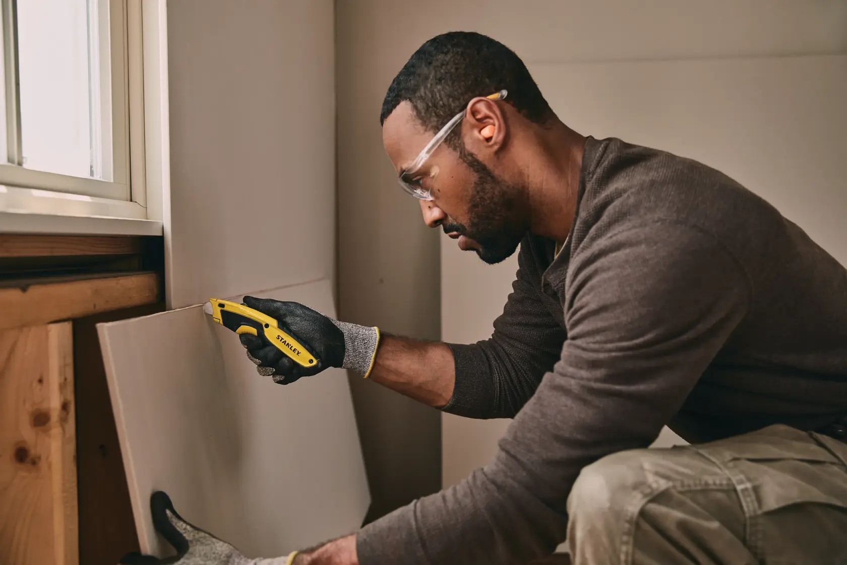 Estilete STANLEY STHT10479 segurado por uma mão com luva cortando drywall em um ambiente interno.
