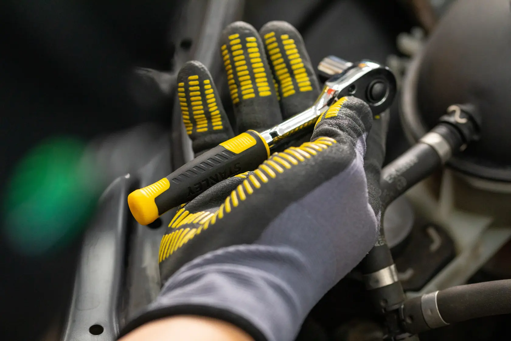 Chave catraca STANLEY STMT82832-LA com cabo preto e amarelo, segurada por mão com luva em cenário de reparo automotivo.
